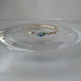 Laurie Fleming Jewellery's "Sapphire Water Lily Ring," a delicate solid gold stacking ring with blue-green teal sapphires scattered along the band. The ring is placed on a glass dish that makes it look like it's surrounded by water ripples. The background is a medium grey.