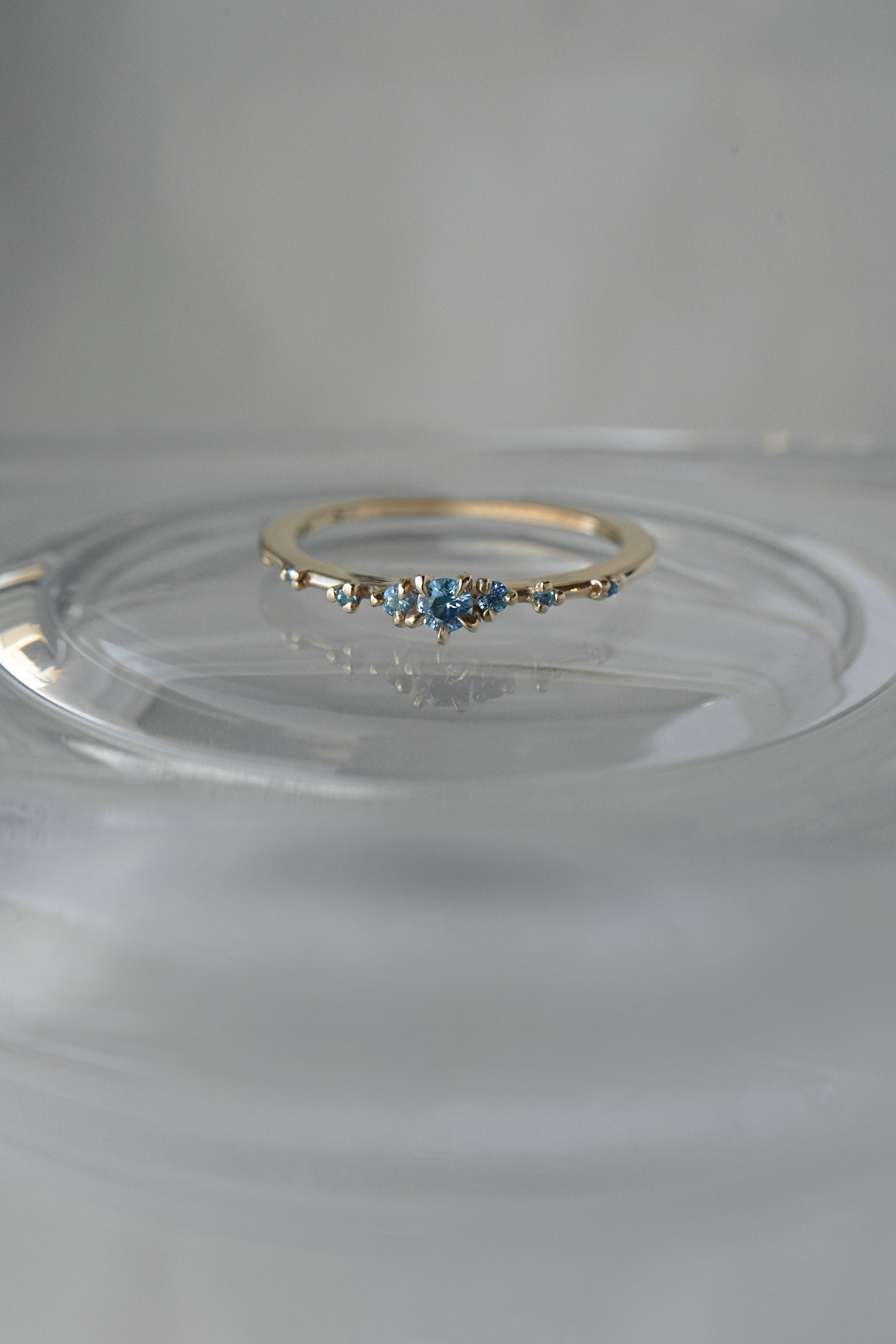 Laurie Fleming Jewellery's "Sapphire Water Lily Ring," a delicate solid gold stacking ring with blue-green teal sapphires scattered along the band. The ring is placed on a glass dish that makes it look like it's surrounded by water ripples. The background is a medium grey.