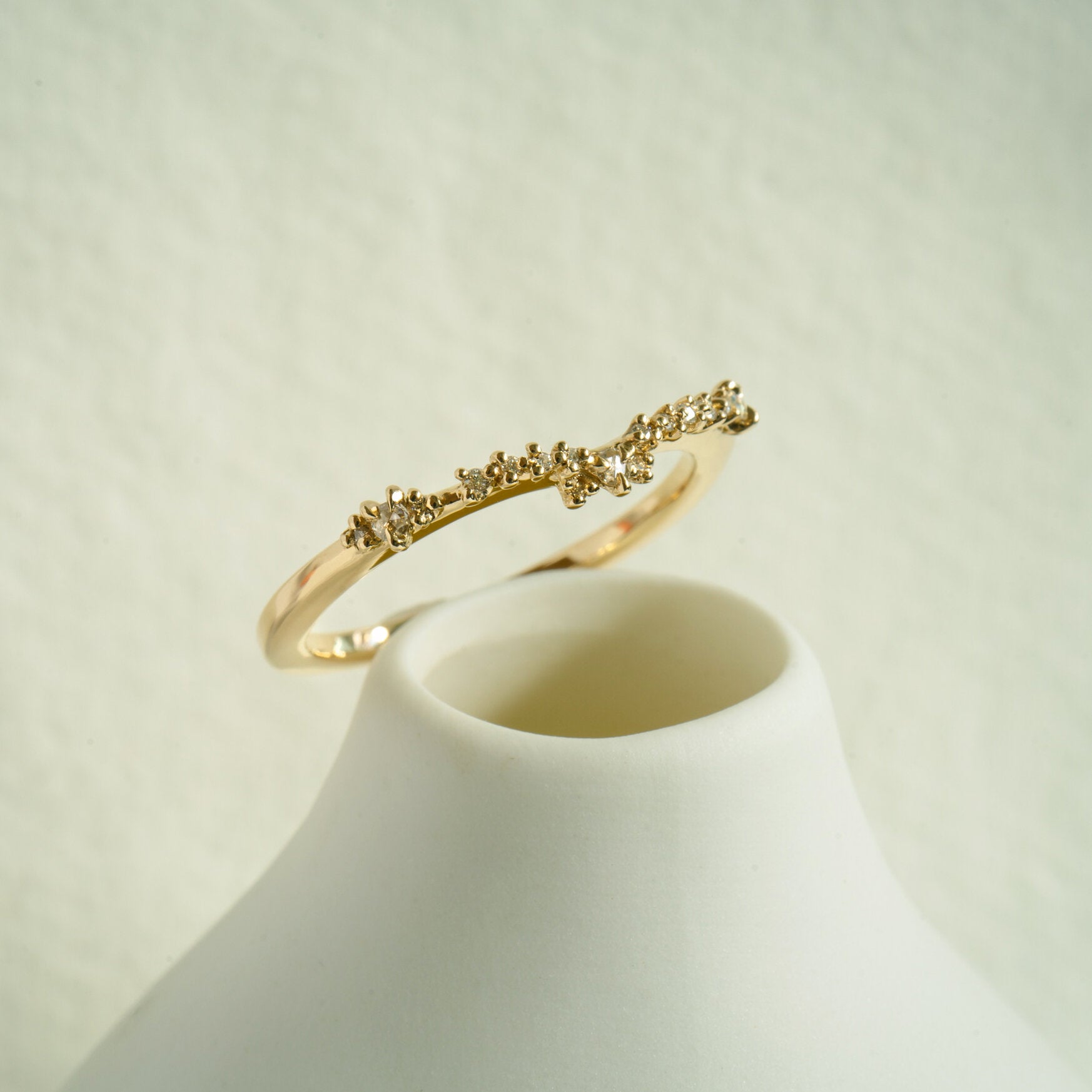 The Sylph Band ring by Laurie Fleming Jewellery, which features a subtly curved band to fit contour around another ring. The band features round brilliant and rose cut diamonds. It is resting at the mouth of a white ceramic vase.